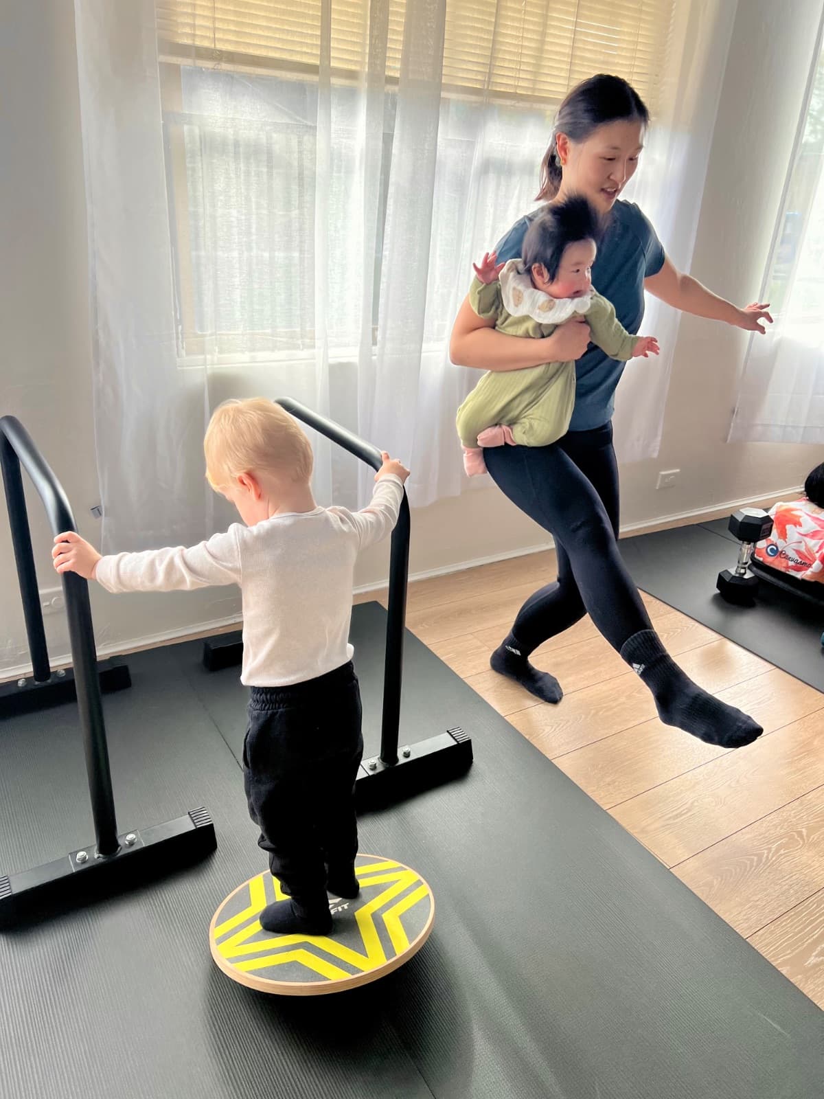 Mum holding baby while toddler plays on balance board during circuit class